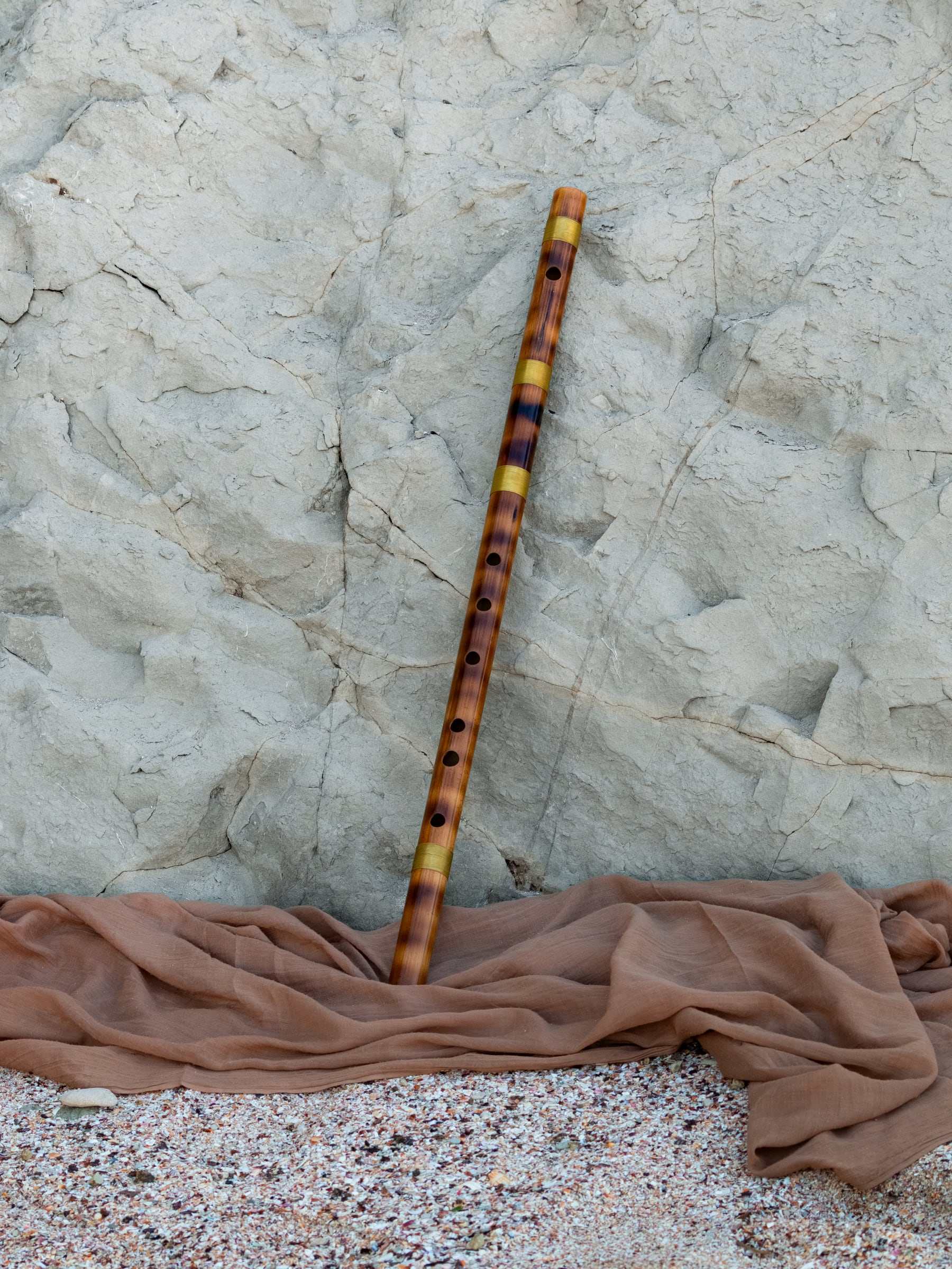 A bamboo bansuri with gold-coloured rings leaning against pale rock on a terracotta cloth