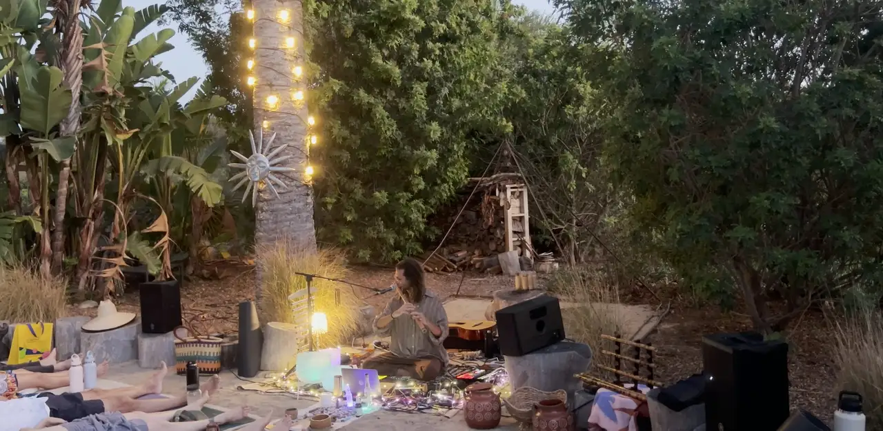 Evening sound journey in a garden: Federico plays the bansuri under a palm wrapped in string lights while listeners lie on the sand