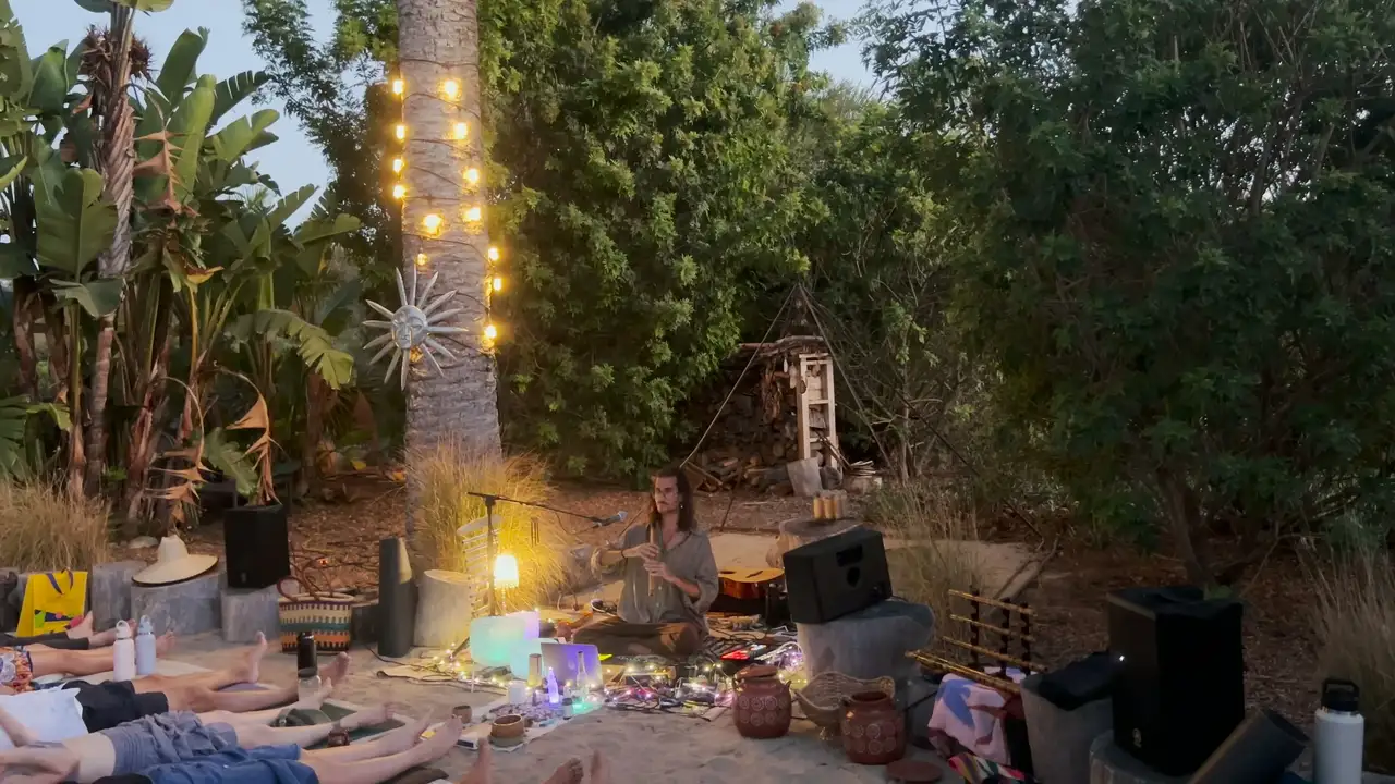 Evening sound journey in a garden: Federico plays the bansuri under a palm wrapped in string lights while listeners lie on the sand